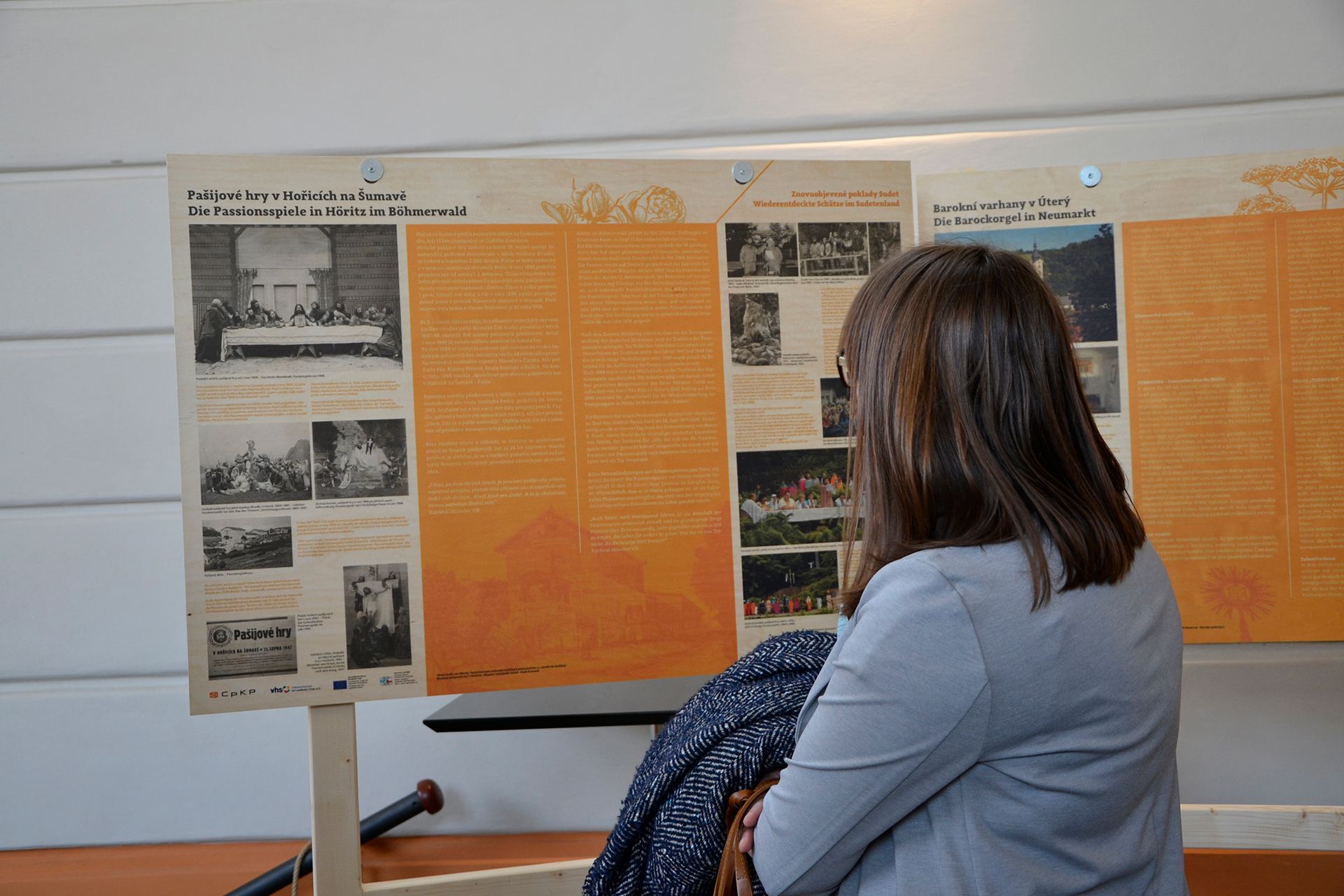 Osoba si prohlíží muzejní expozici s oranžovými panely a historickými fotografiemi na nástěnném displeji