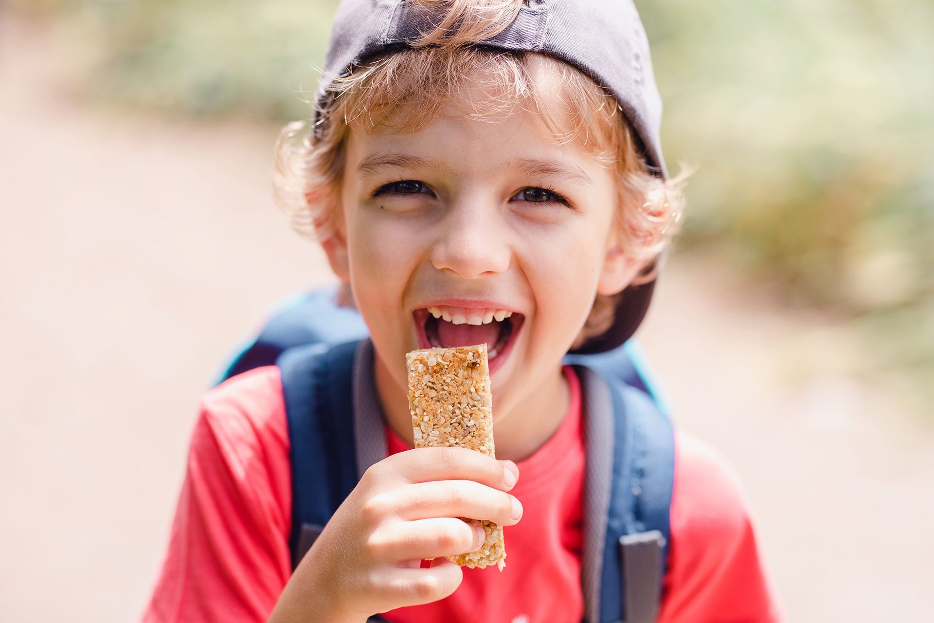 Dítě v červeném tričku a čepici jí venku müsli tyčinku a usmívá se do kamery.