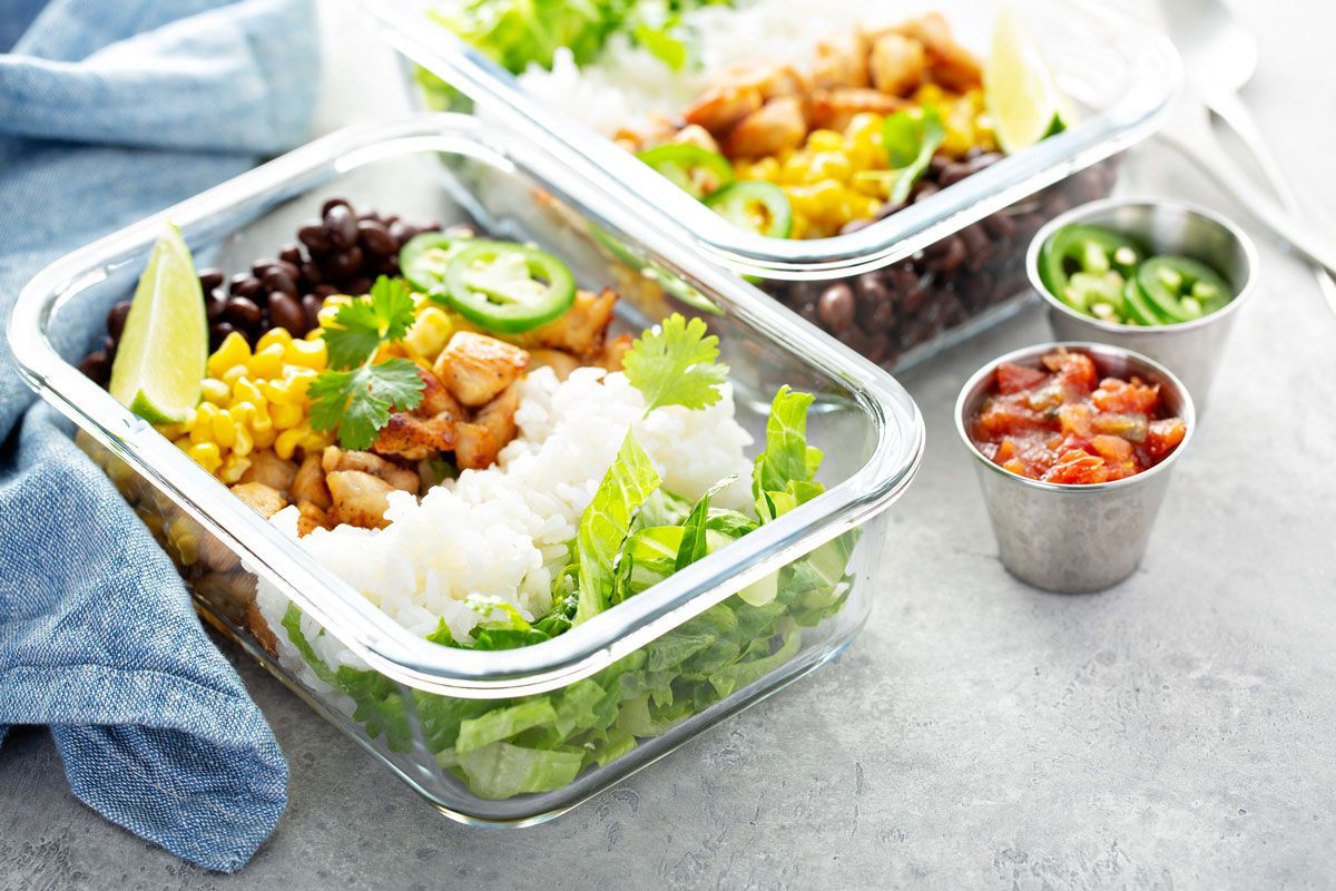 Two glass containers filled with a rice bowl meal, including chicken, black beans, corn, cilantro, and jalapeños. A side of salsa and peppers.