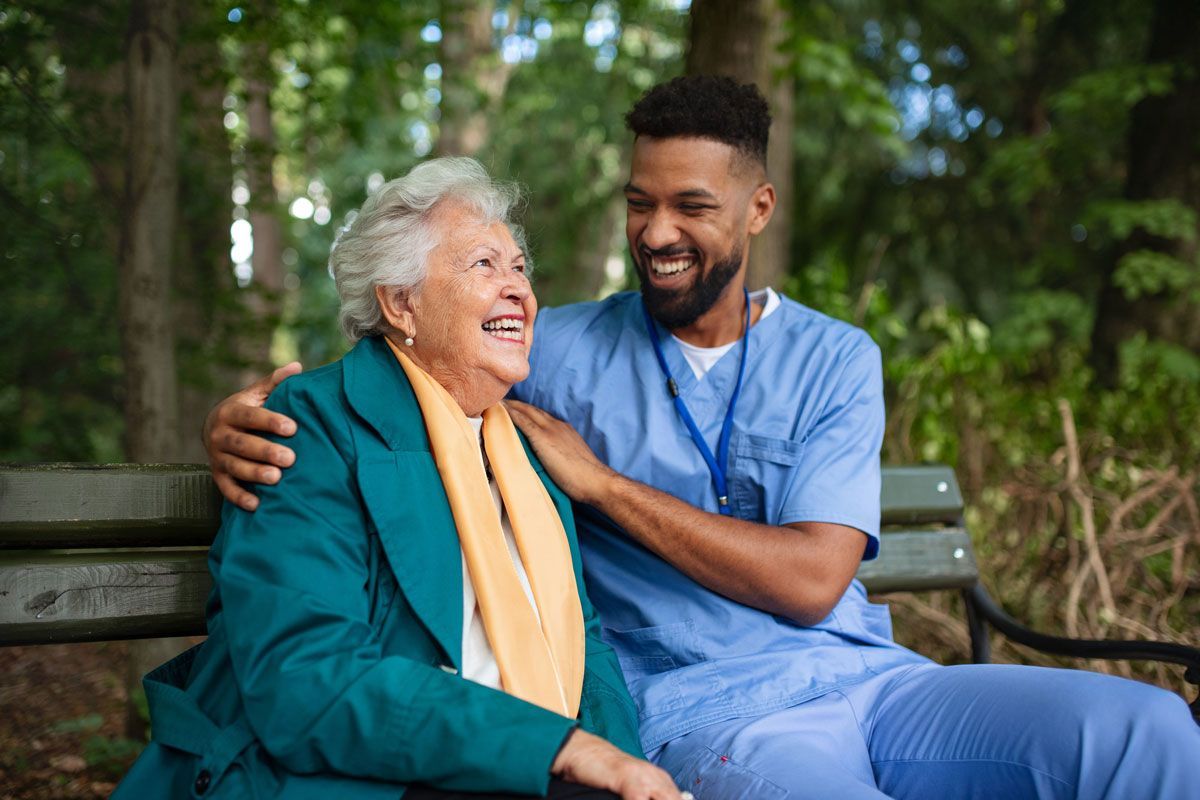 An elderly woman and a smiling male caregiver are seated on a park bench, the man's arm around the woman.