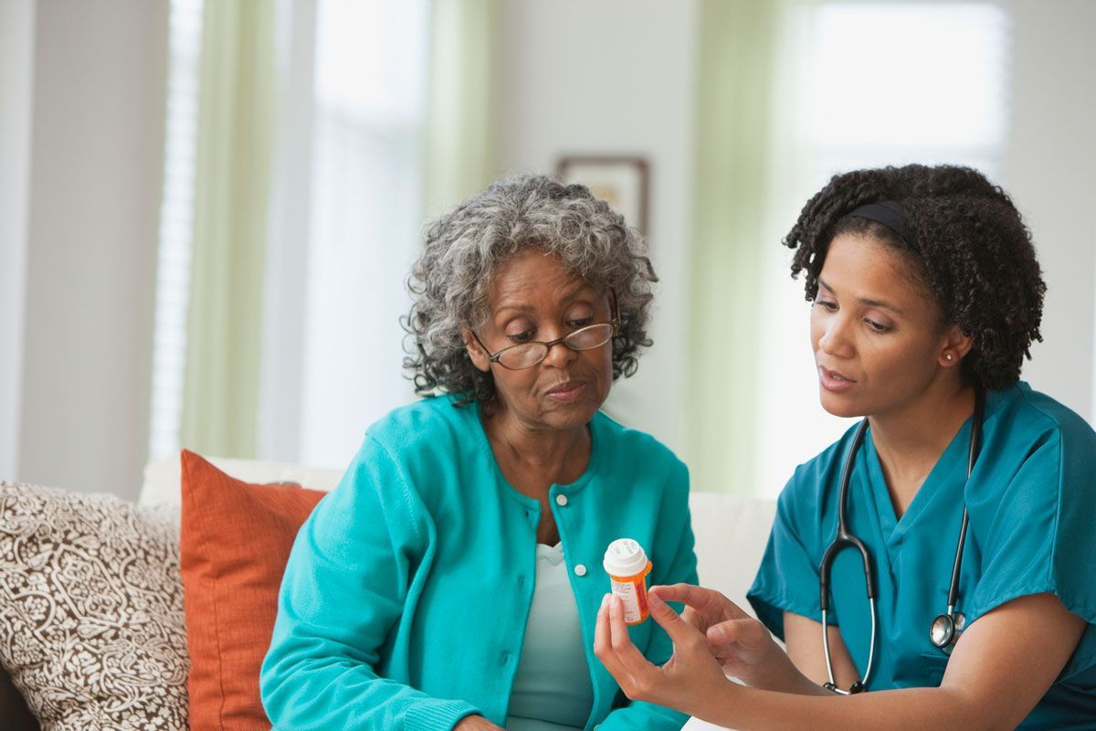A nurse in blue scrubs explains medication to an elderly woman wearing a teal sweater, both seated on a couch.