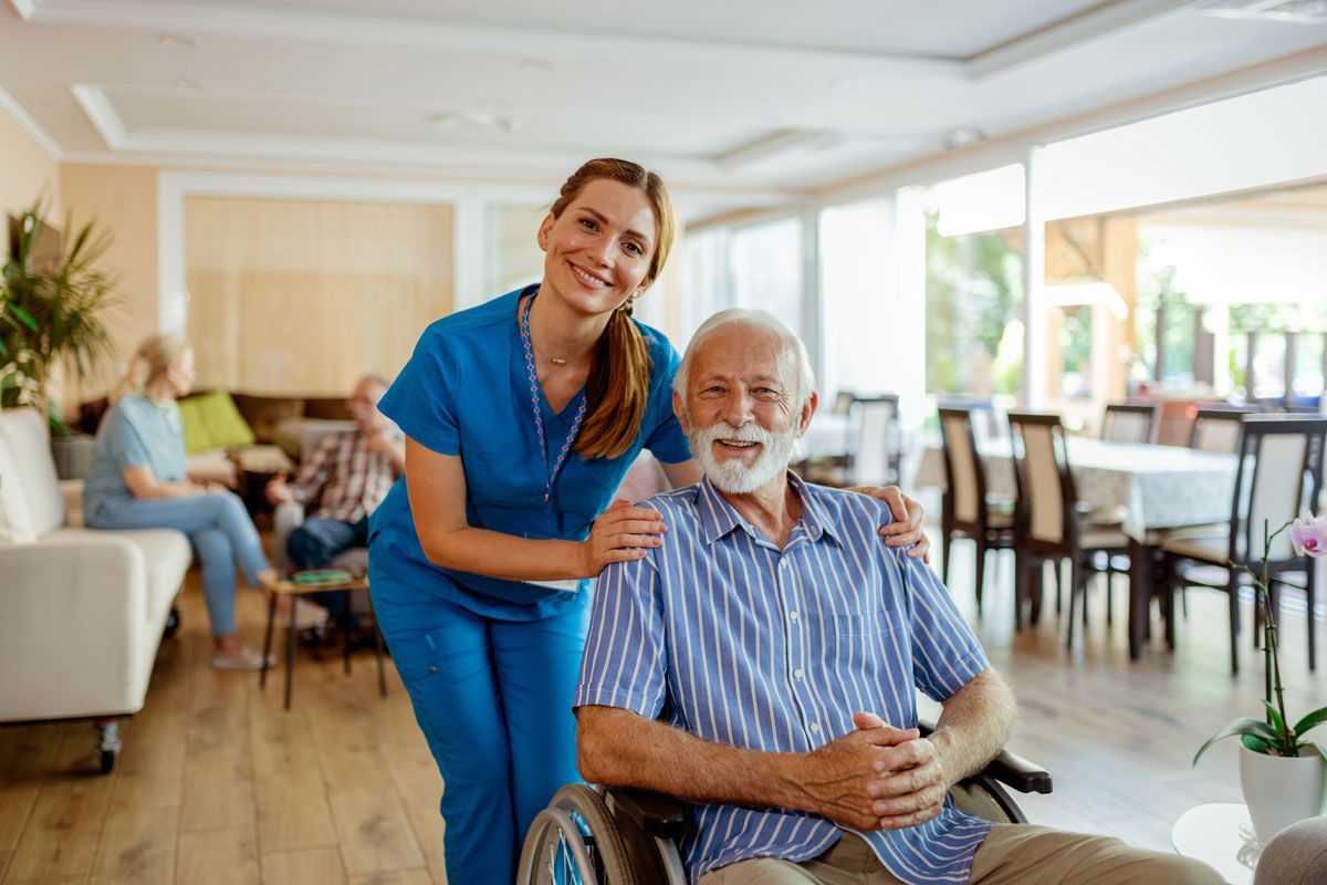 Caregiver in blue scrubs smiles next to an elderly man in a wheelchair, indoors. Other residents are in the background.