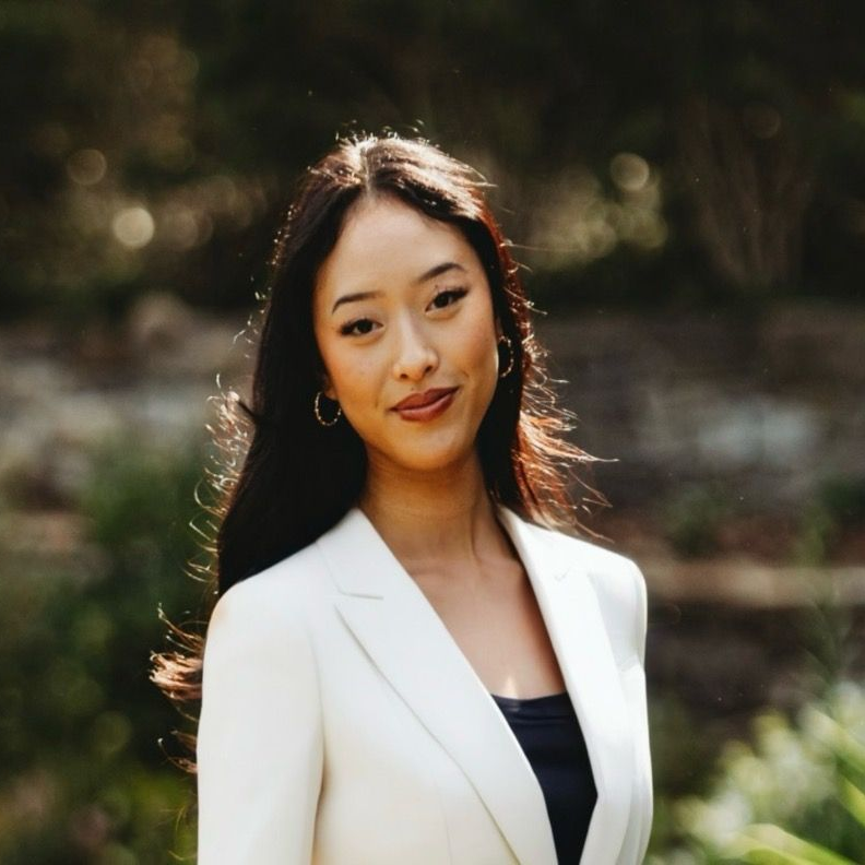 Woman in a white blazer standing outdoors with a soft, blurred green background, smiling gently