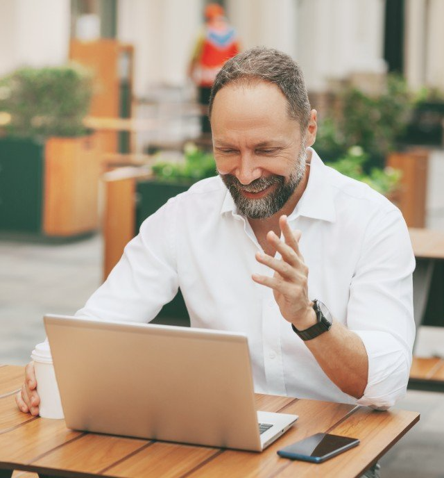 Smiling man in a white shirt waves during a video call on a laptop at an outdoor table