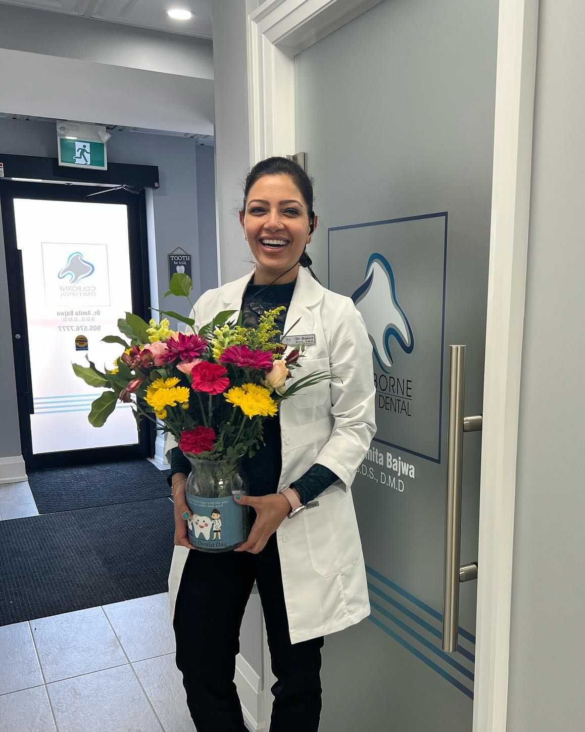 Dr. Amita Bajwa holding flowers, smiling in dental office doorway.