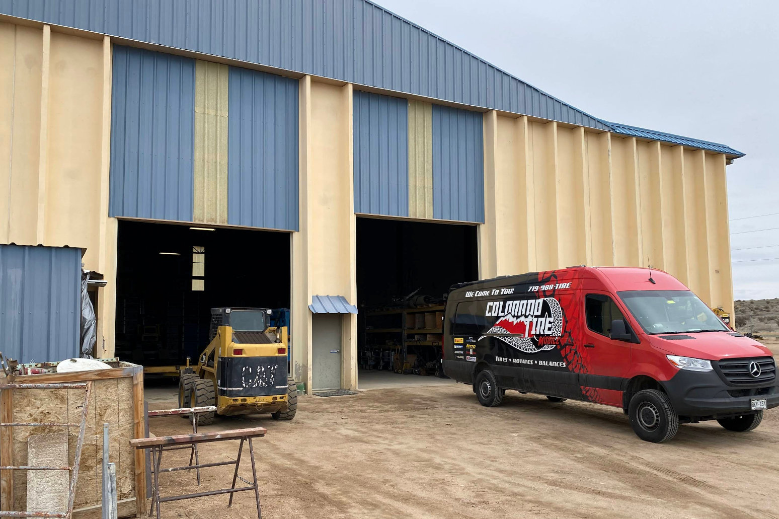 Red van parked outside a blue-and-tan warehouse with open garage doors and equipment inside. | Colorado Tire Auto Care Center