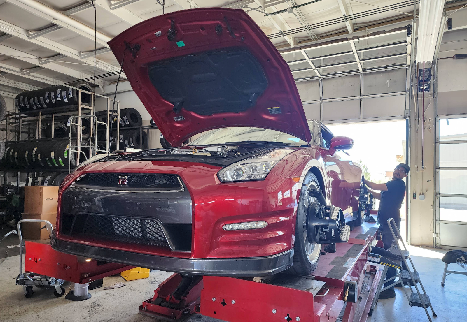 Red sports car on a lift in a garage, hood open, with a mechanic working beside it. | Colorado Tire Auto Care Center