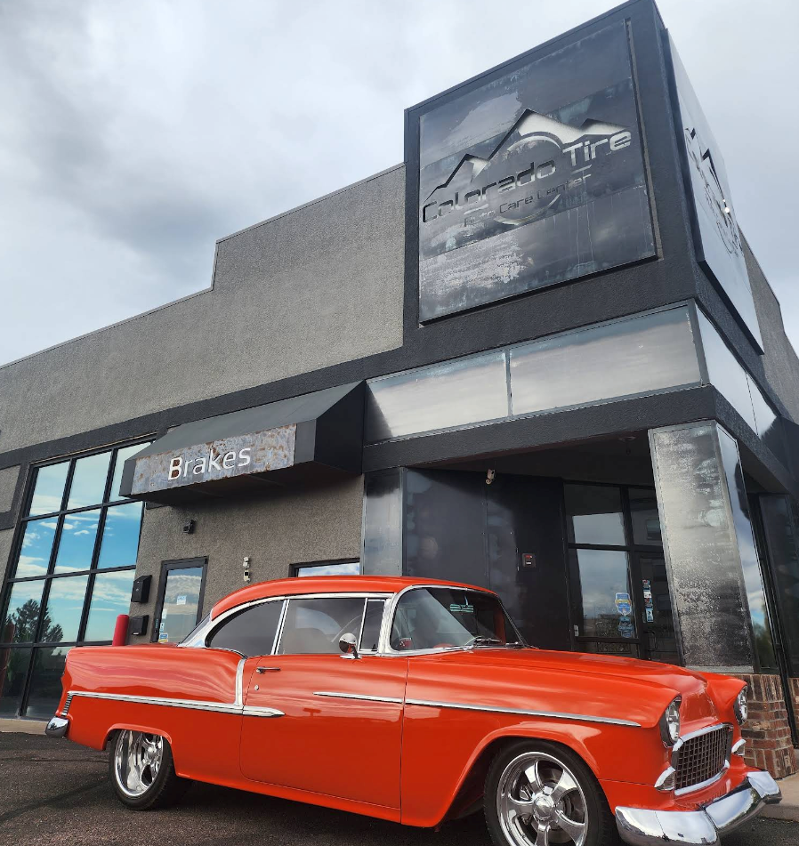 Orange classic car parked outside a modern auto dealership with a large car logo sign | Colorado Tire Auto Care Center