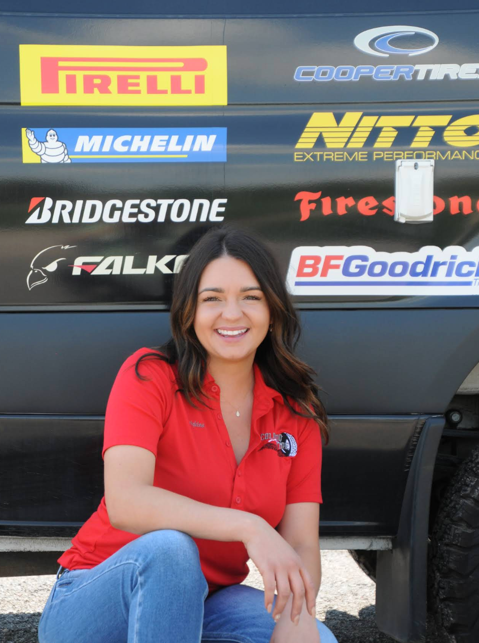 Woman in a red polo shirt sitting in front of a tire brand display, smiling. | Colorado Tire Auto Care Center