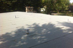 Toit plat avec cheminée et conduits de ventilation, arbres en arrière-plan, zone ombragée sur le toit.