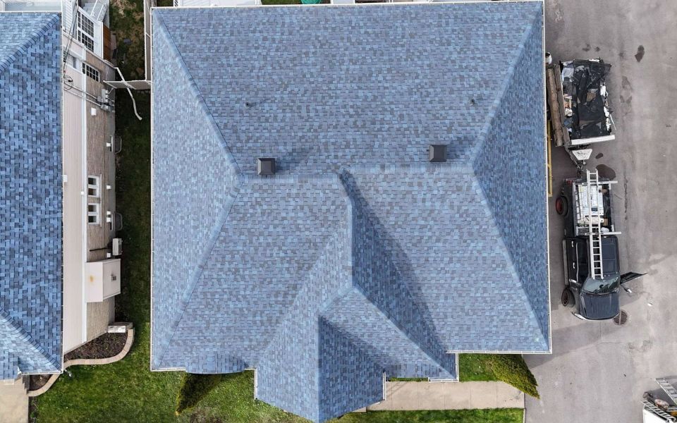 Vue aérienne d'un toit en bardeaux bleu-gris sur une maison, avec deux cheminées, adjacente à un véhicule et à un autre bâtiment.