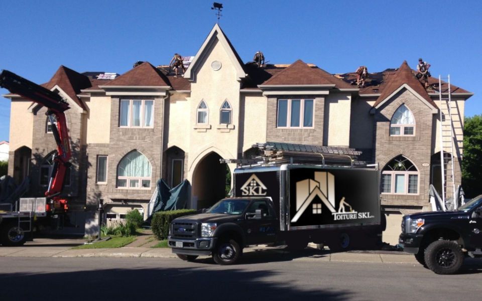 Des camions portant le logo d'une entreprise de couverture sont stationnés devant une maison. Des ouvriers travaillent sur le toit, une grue soulève des matériaux. Ciel dégagé et ensoleillé.