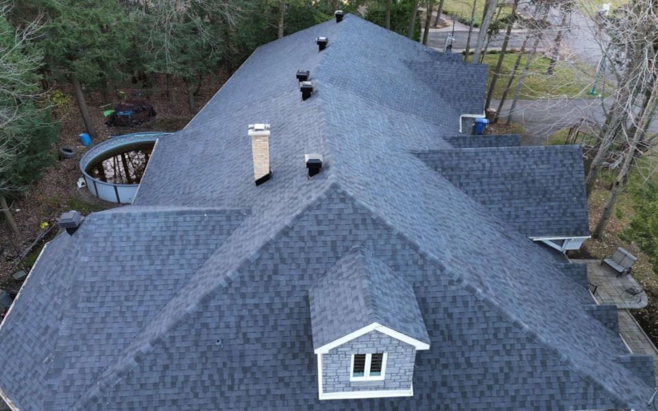 Vue aérienne d'un toit en bardeaux gris foncé avec cheminée et lucarne sur une maison entourée d'arbres.