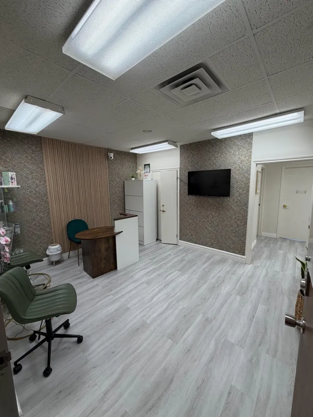 Empty office room with desks, chairs, wall-mounted TV, and bright fluorescent ceiling lights.