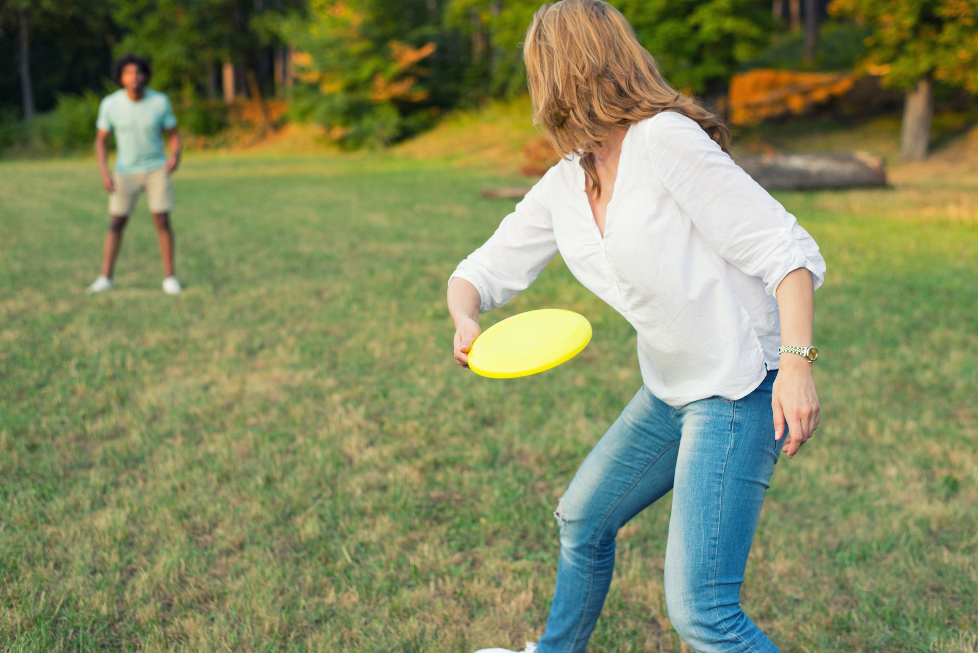 Woman throwing a yellow frisbee in a park towards a person standing in the distance.