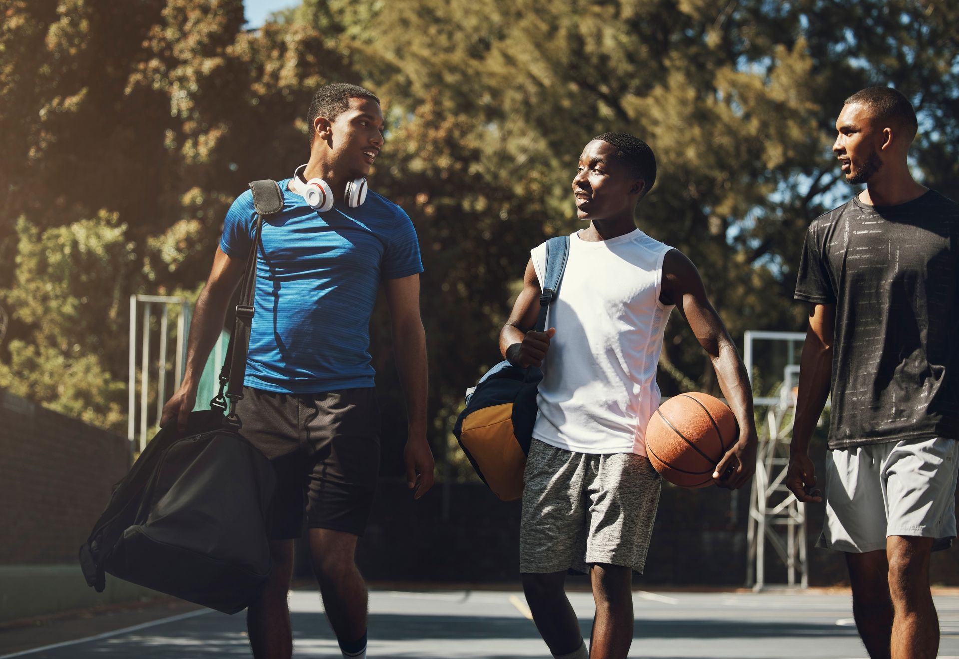 Three people walking on a basketball court, carrying bags and a ball, talking.