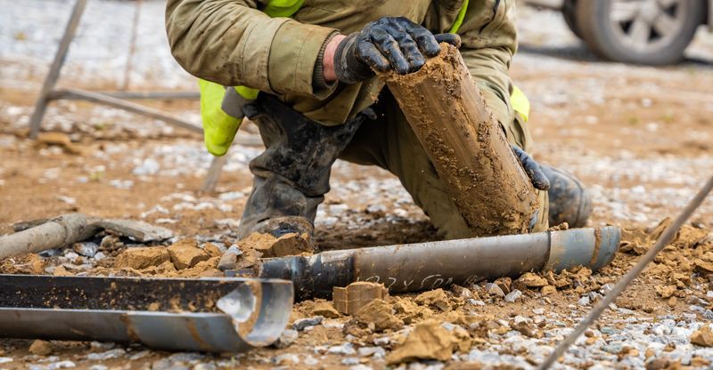 Geological Engineer kneeling, examining a soil core sample, extracted from a drilling pipe on a dirt construction site. The Basics of Geotechnical Engineering: Principles and Practices