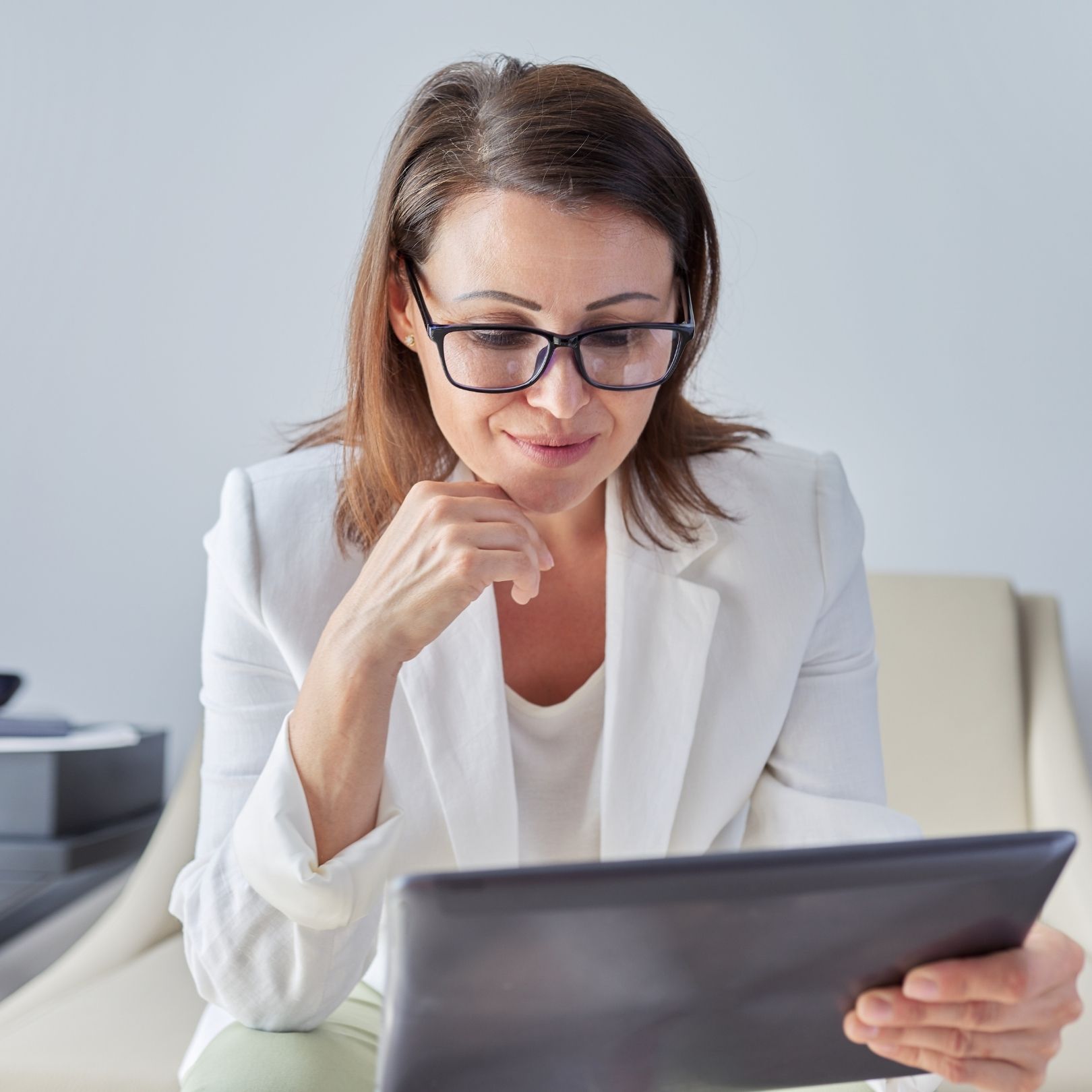 Woman in a white blazer reading a tablet at a desk, with a thoughtful expression.
