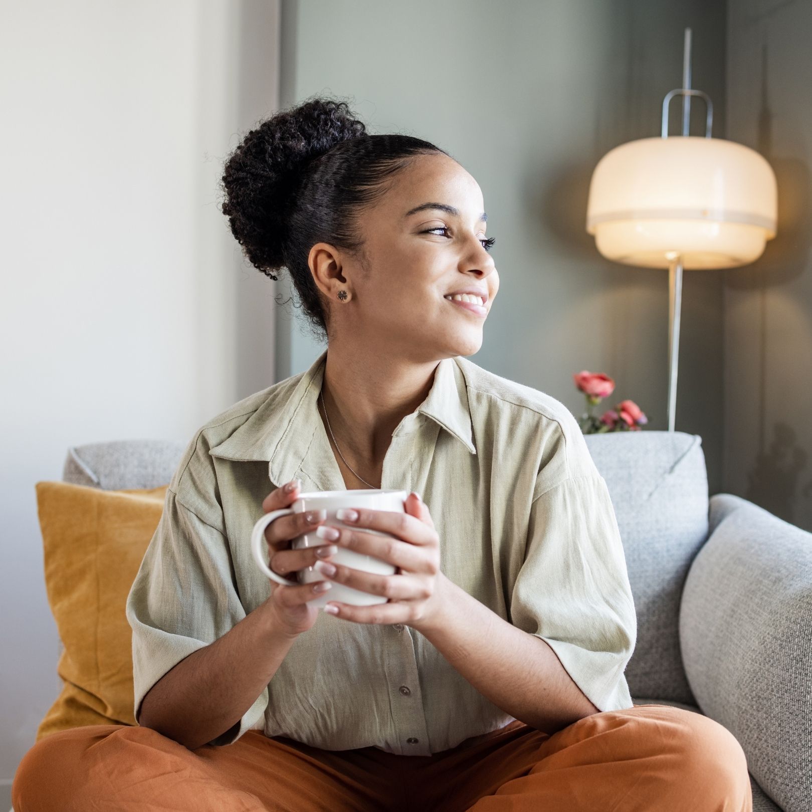 Woman sitting on a couch holding a mug, looking out a window in a cozy living room.