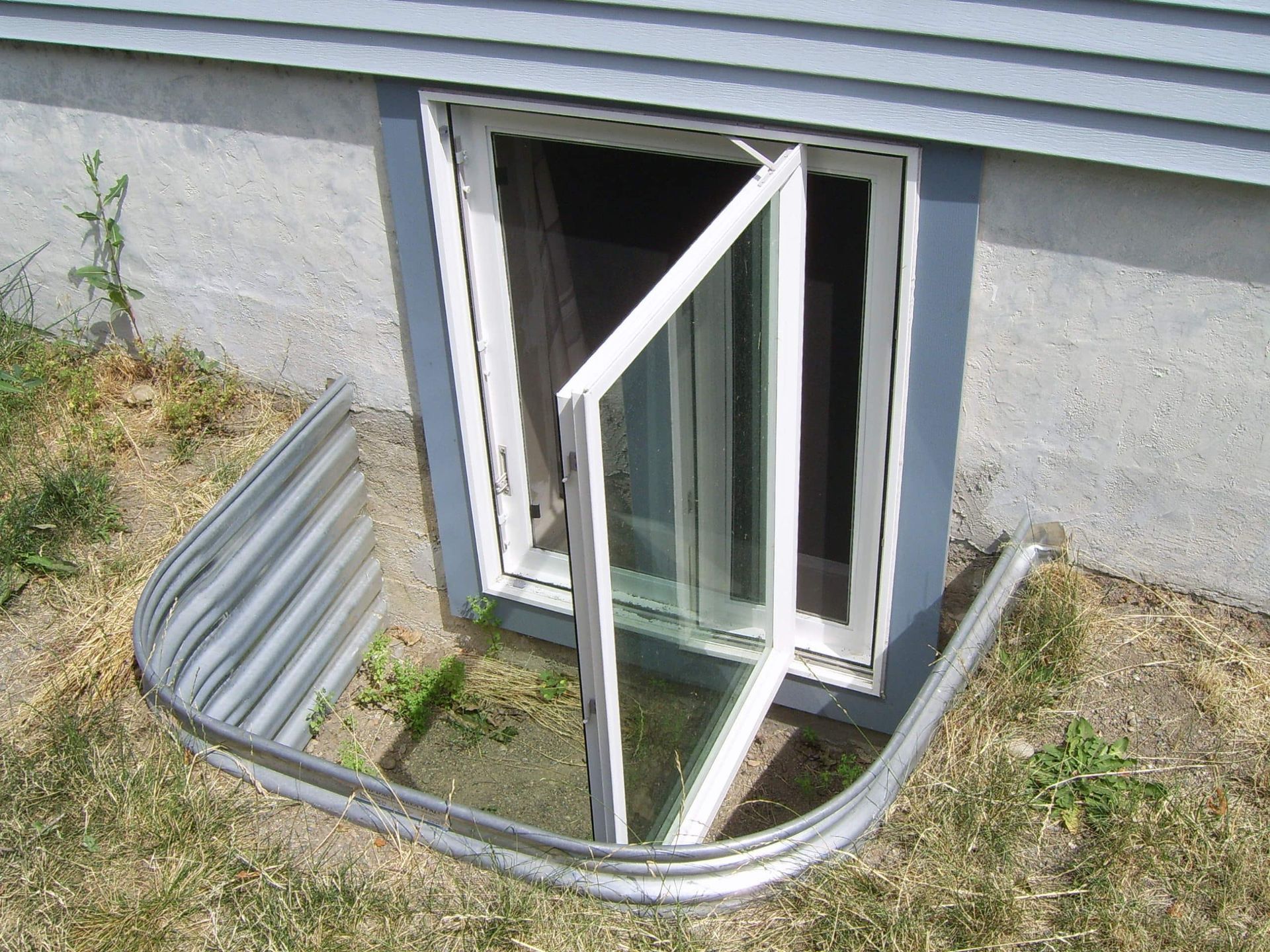 Basement window with open door, silver well, grass, gray siding.