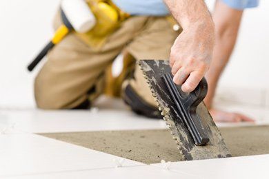 A construction worker kneeling, applying mortar to the floor with a notched trowel, preparing to lay tile.