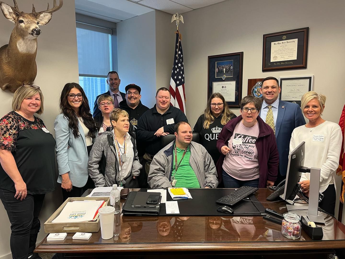 Group photo in an office. People smiling and posing around a desk with an American flag in the background.