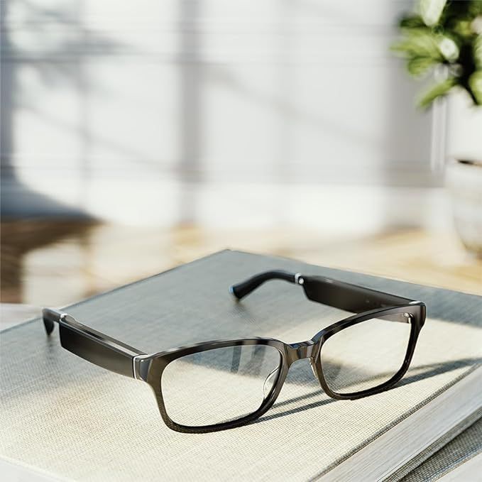 Smart glasses resting on a book. Black frames, clear lenses, sunny background.