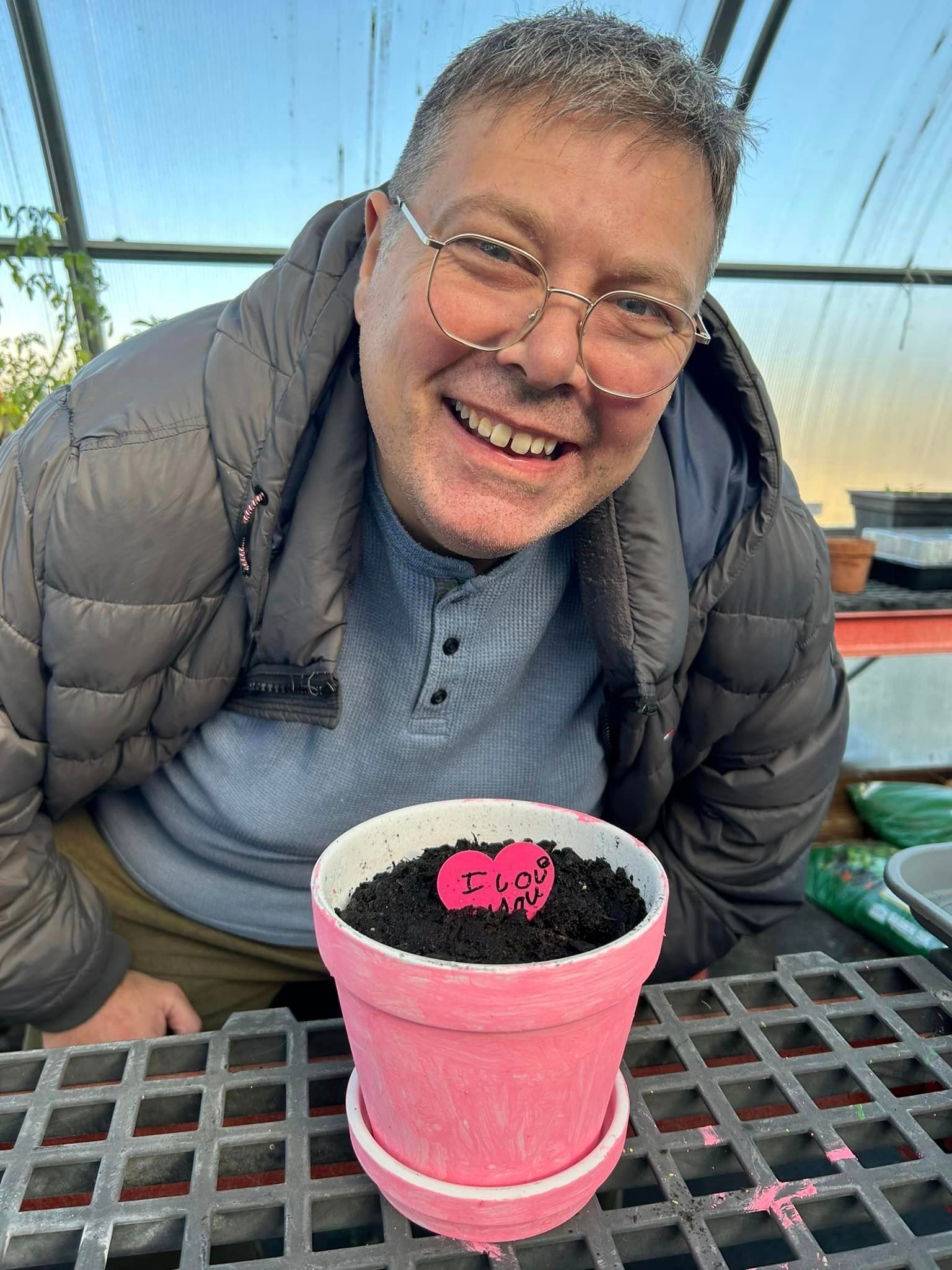 Man smiling, holding pink pot with soil, inside greenhouse.