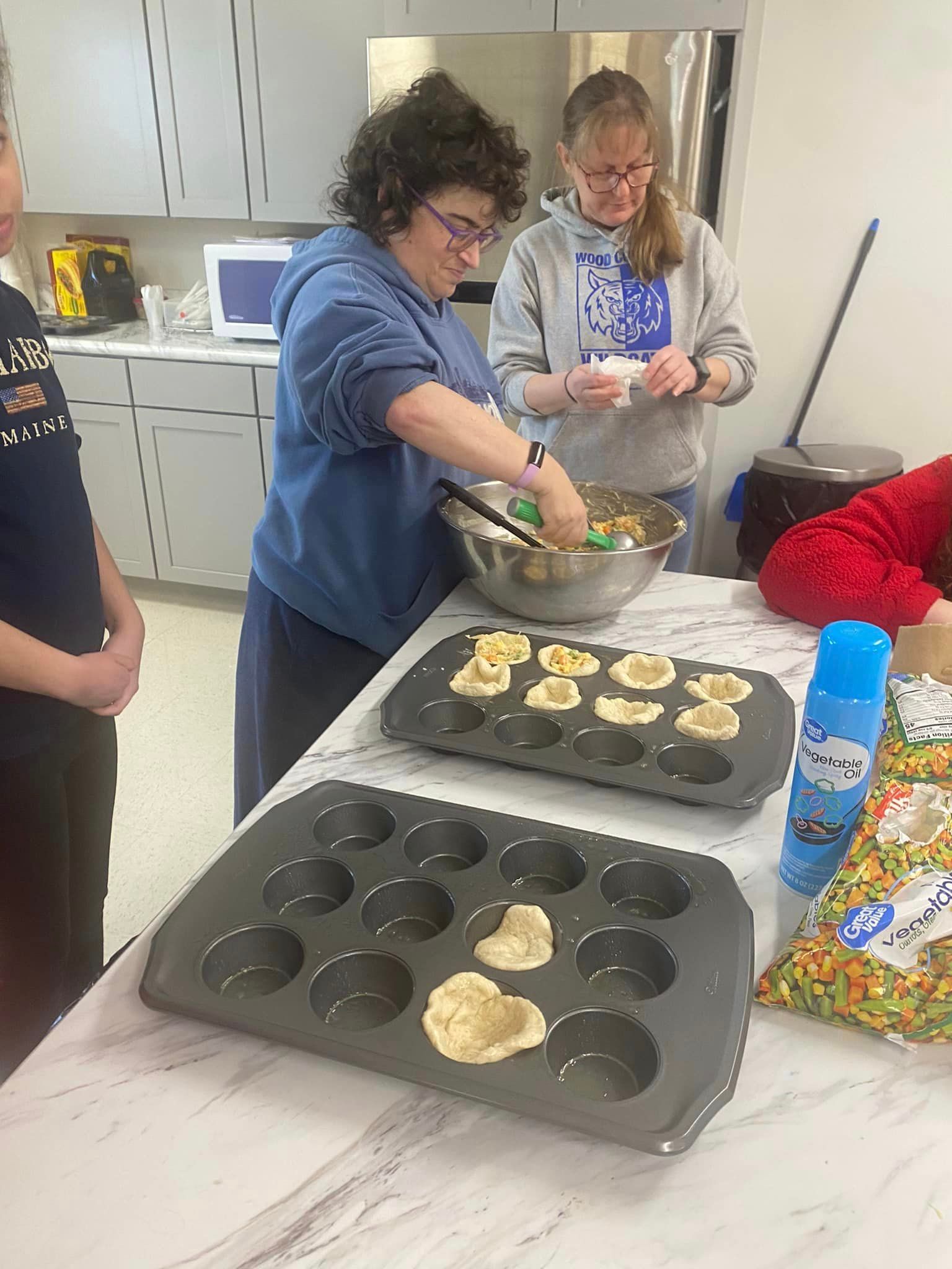 Two women in a kitchen making baked goods. One scoops batter into muffin tins.