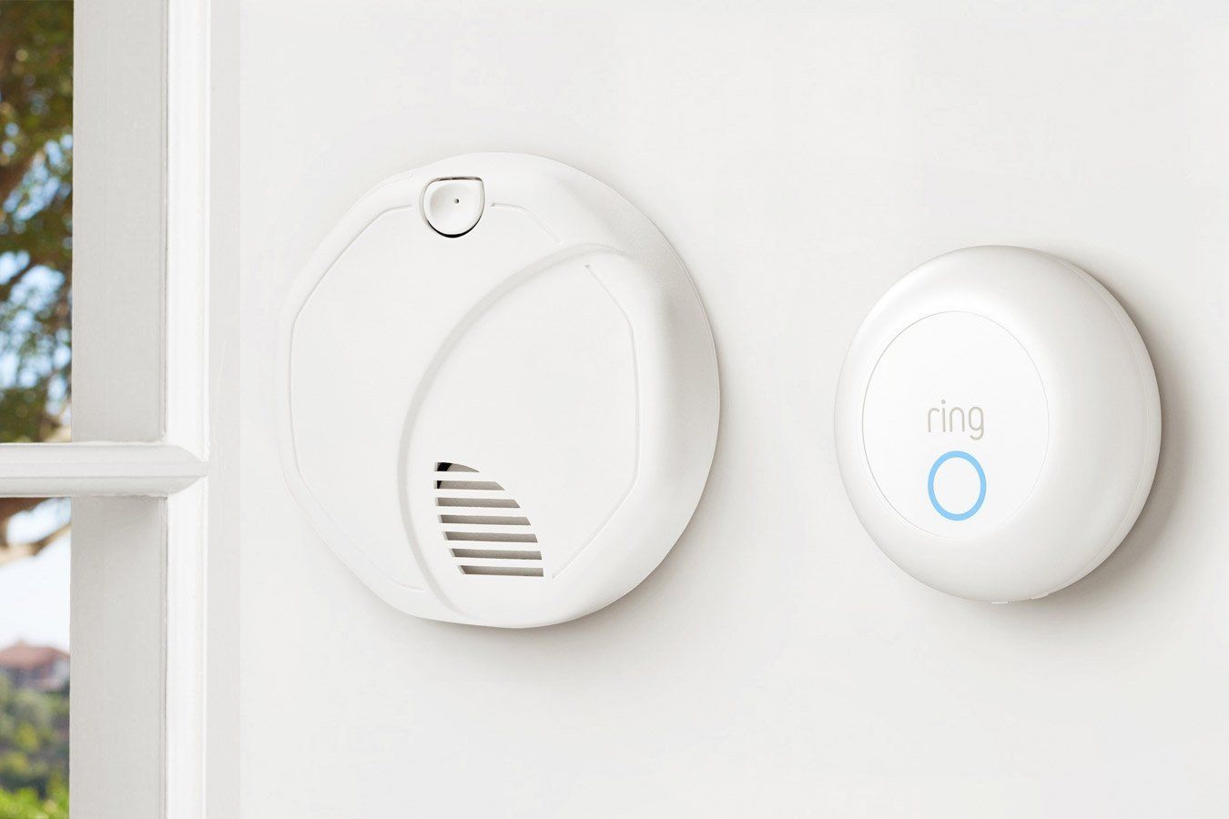 White smoke detector and Ring security device on a wall near a window.