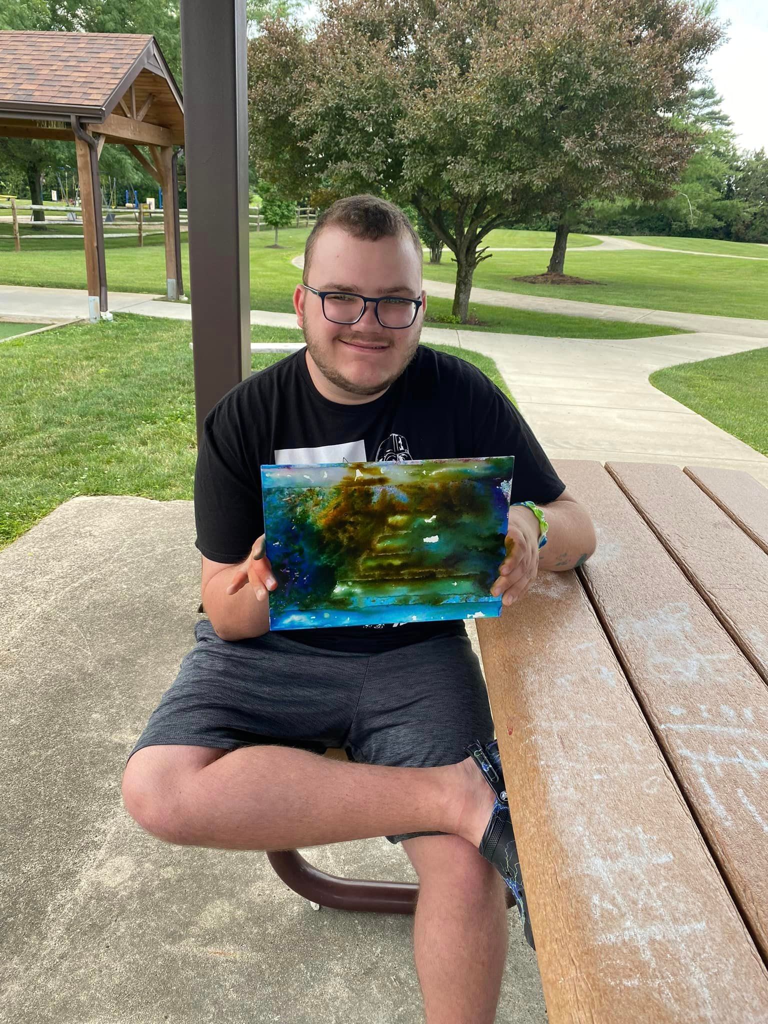 Man with glasses, sitting at a picnic table, holding a colorful piece of art, smiling in a park setting.