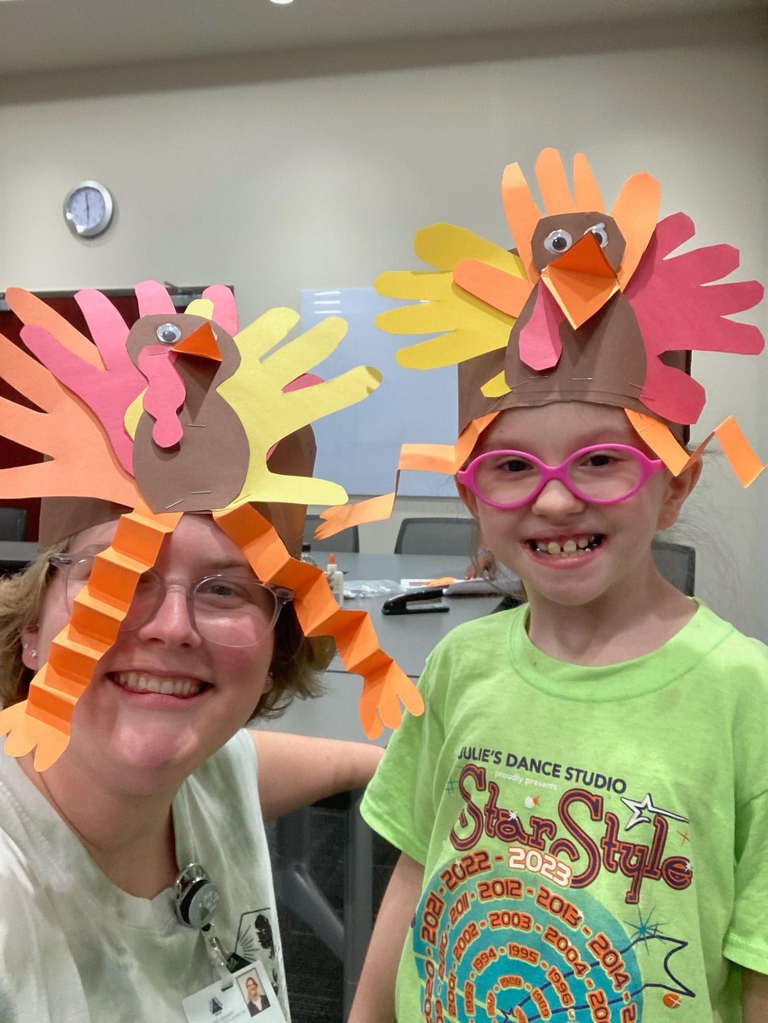 Two people with turkey handprint hats, smiling at the camera. Indoors with a clock on the wall.