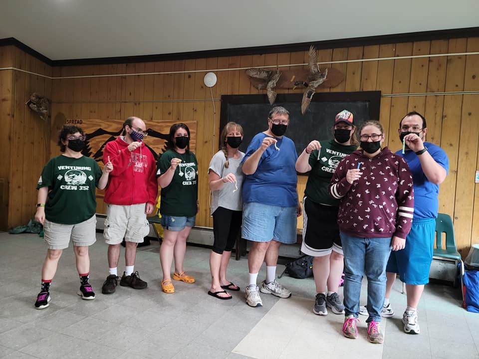 Group of masked people holding items in a room with wood paneling. They appear to be posing, some smiling.