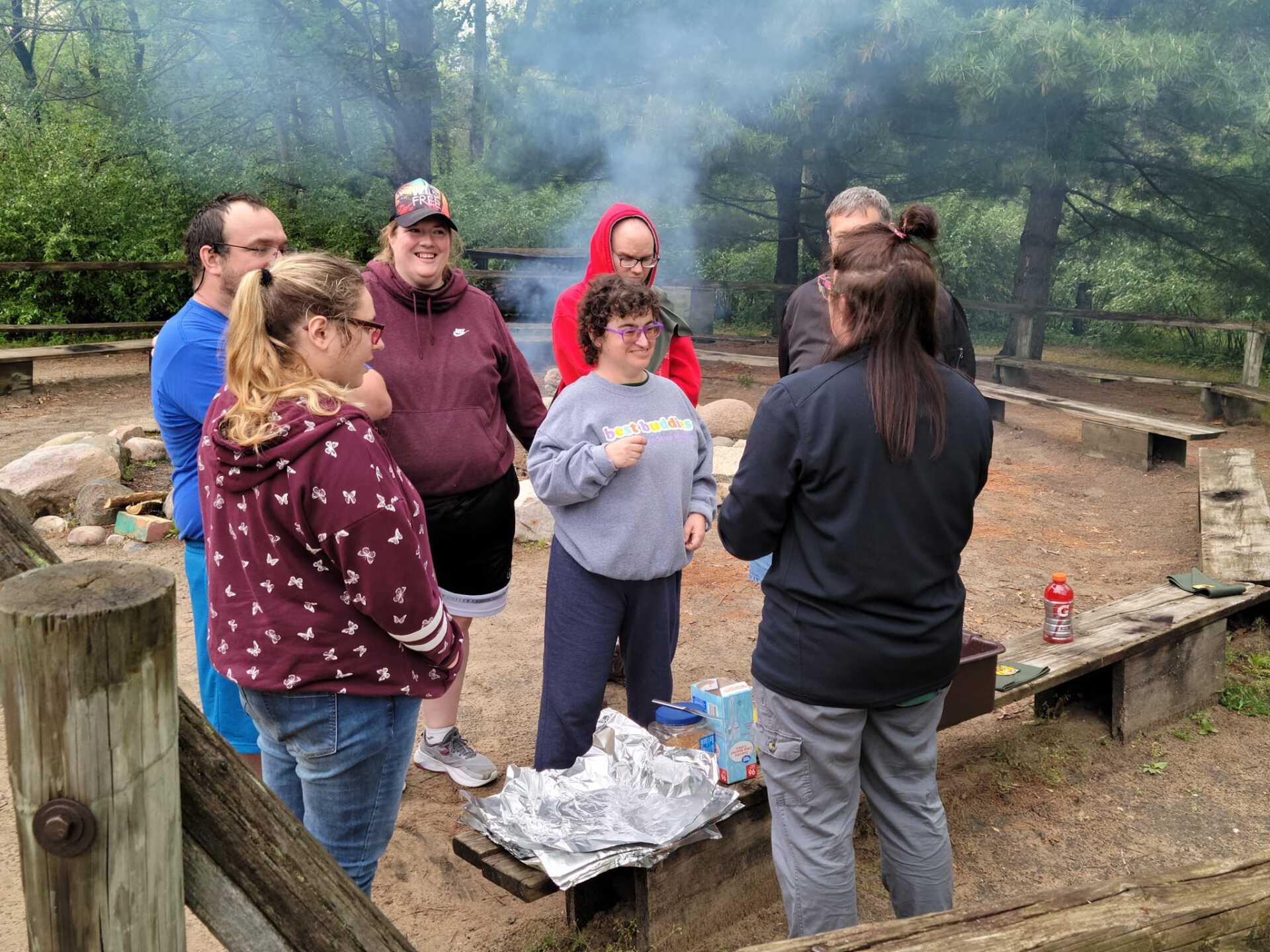 Group of people gathered around a campfire in a wooded area, some are smiling, foil is nearby.