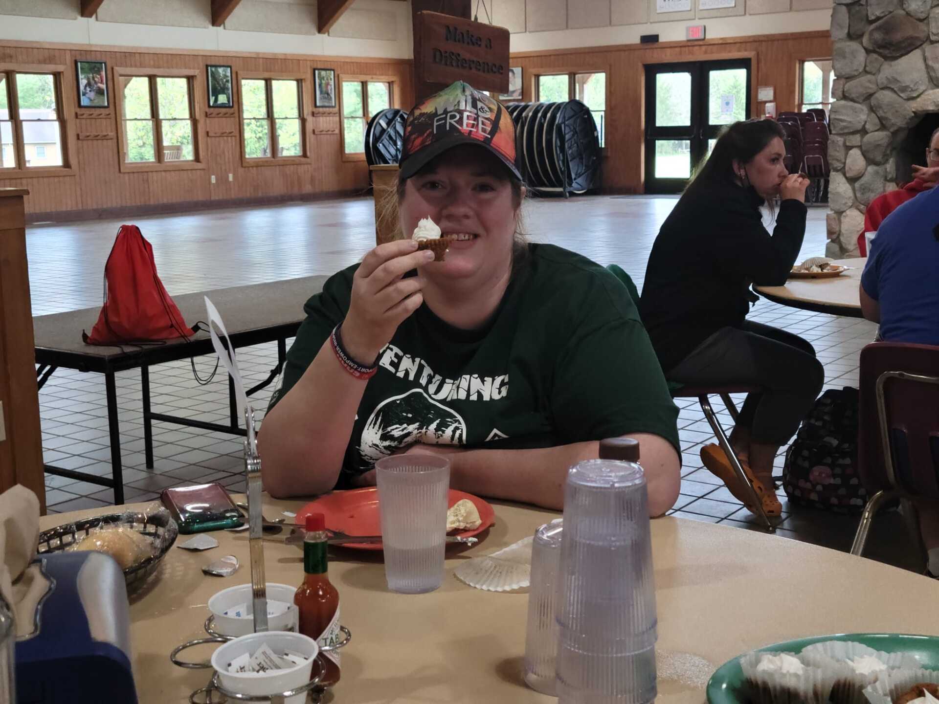 A woman smiling, holding food at a table. Eating in a cabin-style dining area with others.