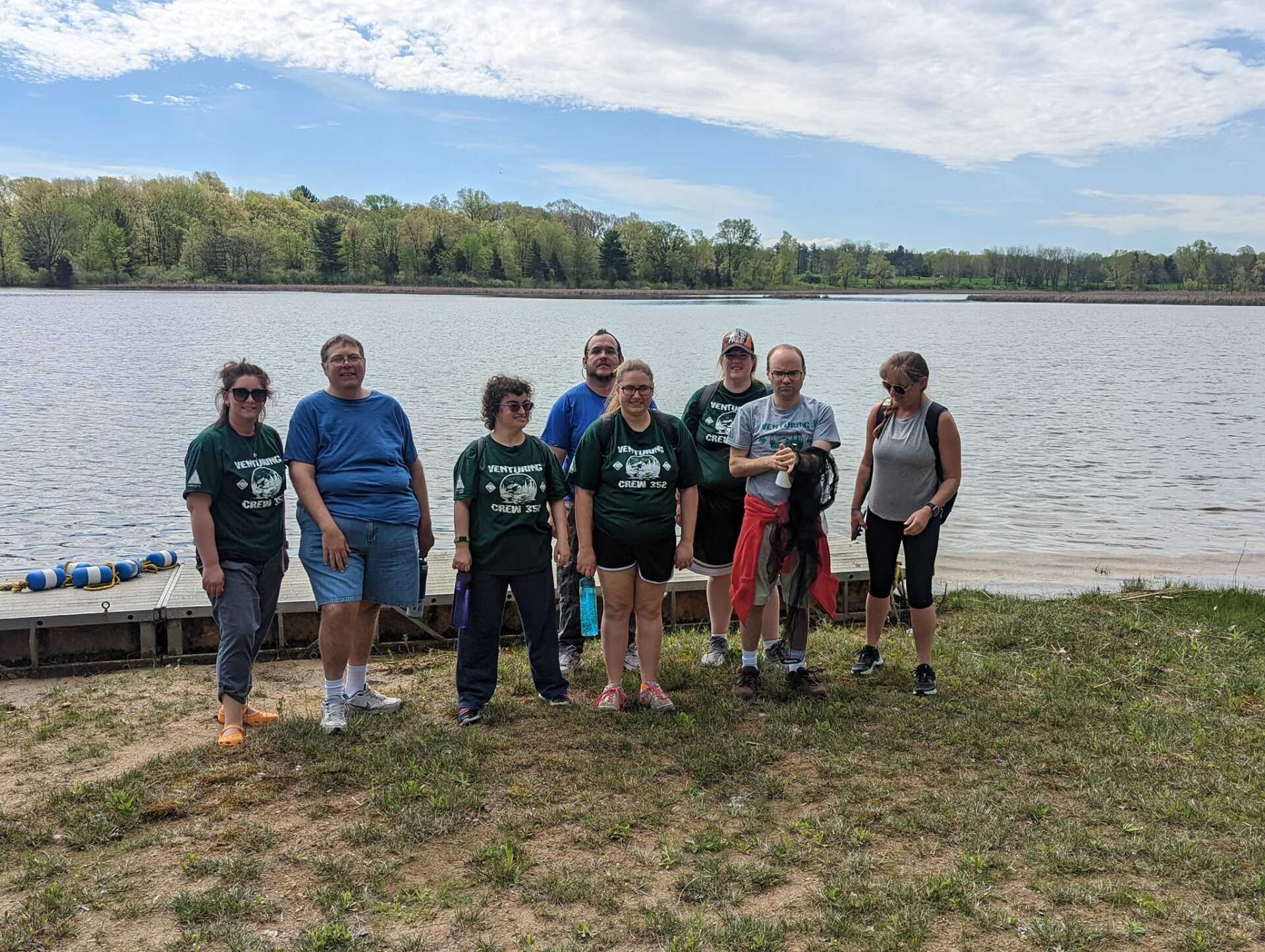 Group of people standing near a lake on a sunny day, some wearing green shirts, looking at the water.