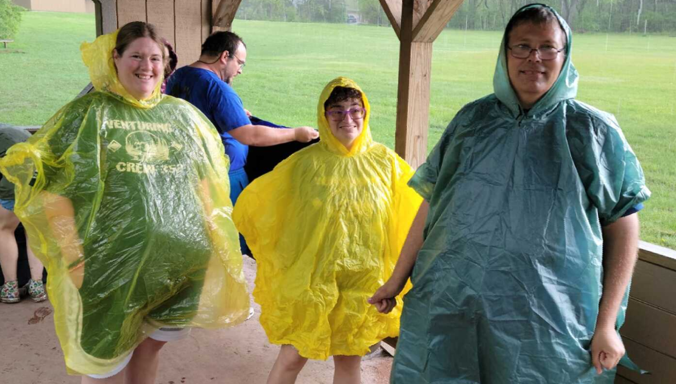 Three people wearing rain ponchos, green, yellow, and blue-green, standing outdoors under a shelter.