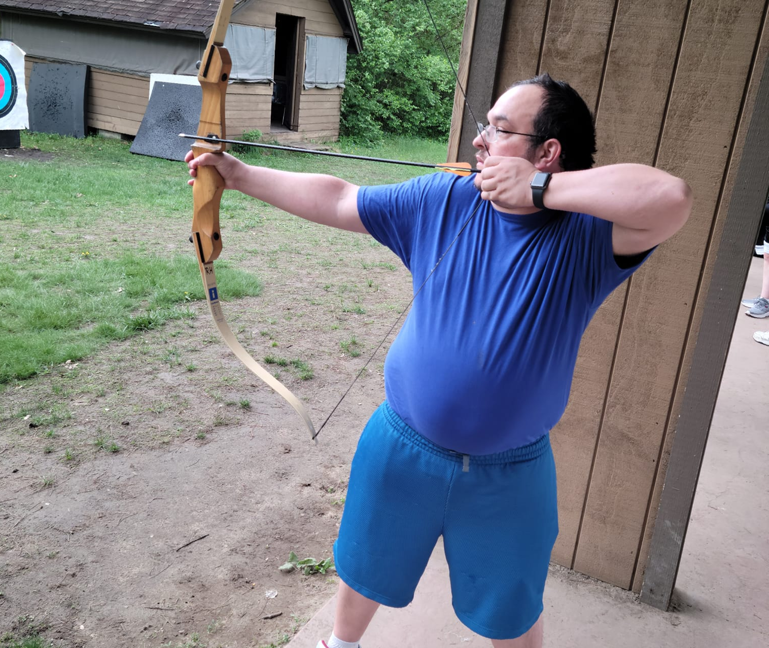 Man in blue shirt and shorts aims a bow and arrow at a target outdoors.