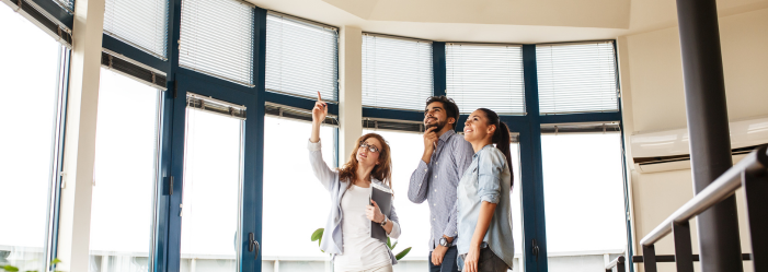 A real estate agent points at the high ceiling to a couple in a modern building with large windows.