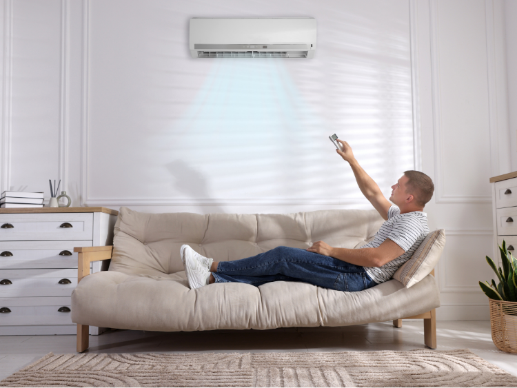 Man on a sofa adjusts air conditioner with remote. Blue air stream visible. Bright room.