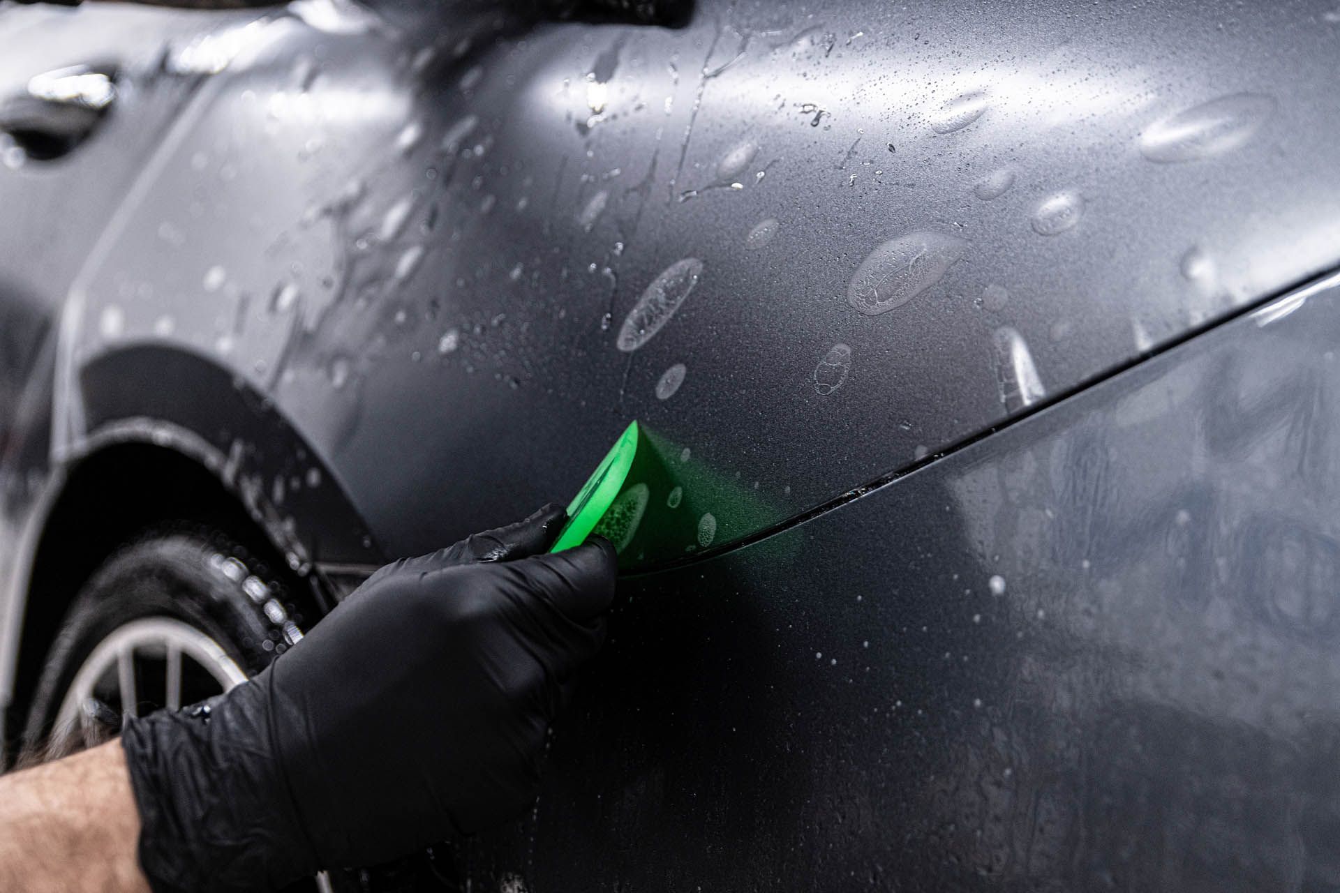 A person in black gloves uses a green tool to apply something to a car's dark side.