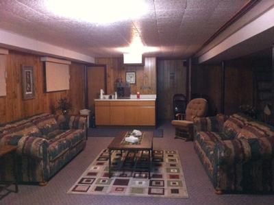 Wood-paneled living room with two sofas, coffee table, rug, and a lit ceiling lamp with a kitchenette at the back
