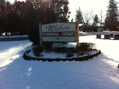 Snowy roadside sign for Macer Funeral Home, surrounded by trees and a low stone border.