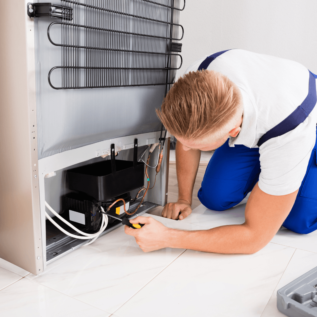 Un hombre está arrodillado en el suelo arreglando un refrigerador.