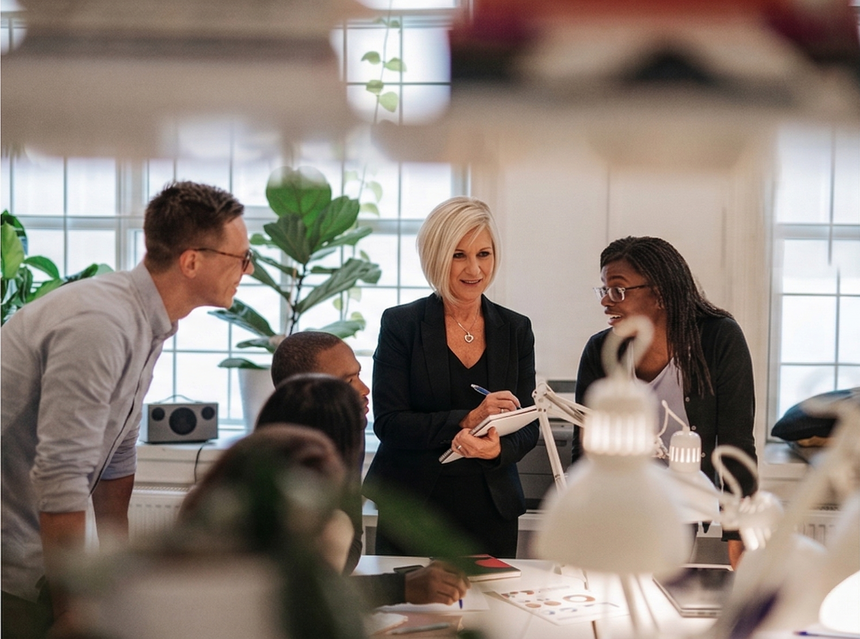 A diverse group of colleagues collaborate at a table in a bright office, with one person writing on a document. A diverse group of colleagues collaborate at a table in a bright office, with one person writing on a document.