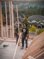 Two construction workers building a wooden frame on a rooftop overlooking houses and trees. Two construction workers building a wooden frame on a rooftop overlooking houses and trees.
