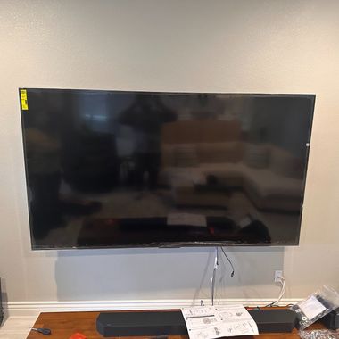 A large black flat-screen television is mounted on a light gray wall above a dark soundbar on a wooden surface.