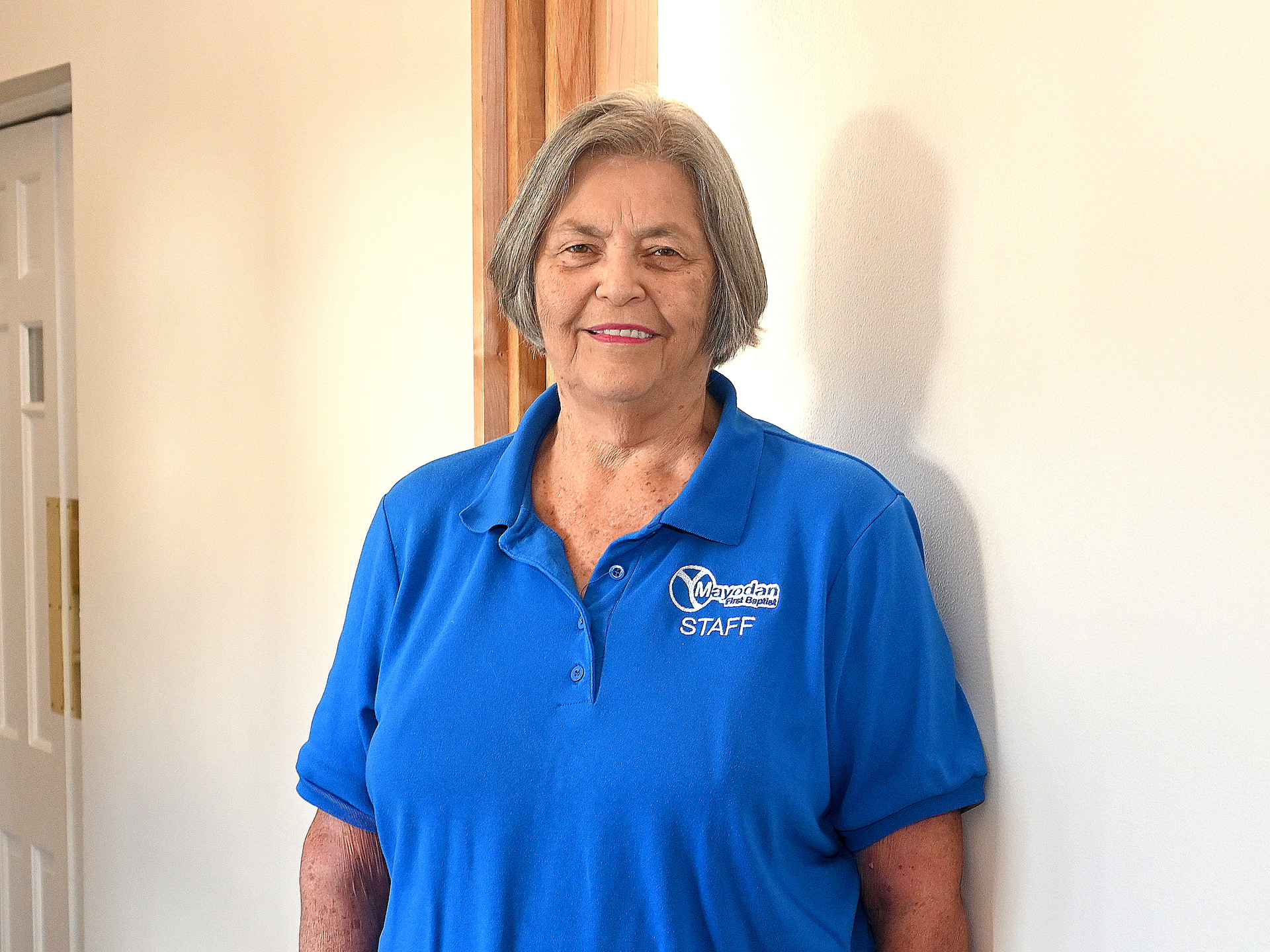 A woman in a blue shirt is standing in front of a white wall.
