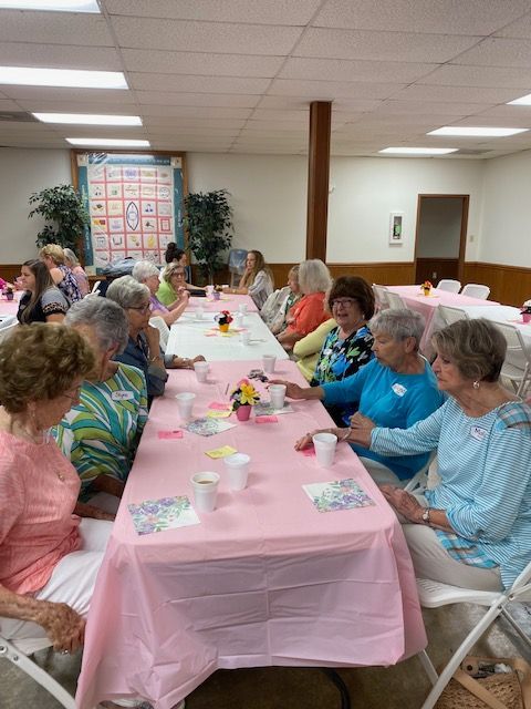 A group of older women are sitting at long tables with pink tablecloths.