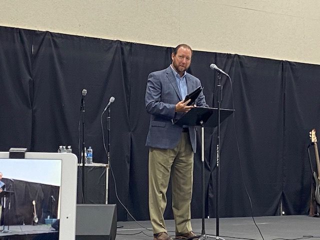 A man is standing at a podium holding a book and speaking into a microphone.